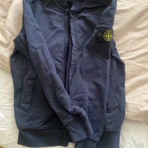 Mörkblå cardigan i bomull från Stone Island Junior - Snygg mörkblå bomberjacka från Stone Island Junior med klassisk logga på ärmen. Jackan har dragkedja framtill, två fickor och ribbade muddar. Perfekt för dig som vill ha en stilren och sportig look.