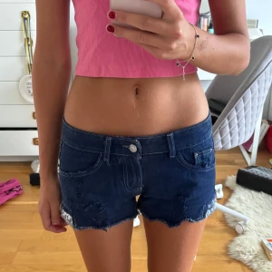 Ultra lowwaist jeansshorts med paljetter - Köpta utomlands! Storlek XS. Bara använda en gång 💕.         Snygga mörkblå jeansshorts med slitna detaljer och råa kanter. Shortsen har paljetter i silver och guld längs bensluten för extra bling. Klassisk femficksmodell med låg midja och cool, avslappnad vibe. Perfekta för sommardagar när du vill sticka ut lite extra.