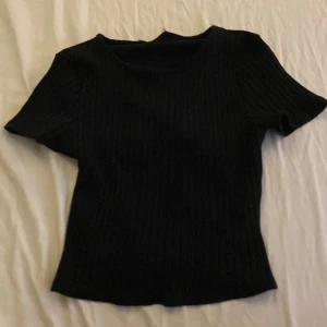 Svart ribbad topp med knyt bak - En svart ribbad topp med kort ärm och rund hals. Toppen har en snygg detalj med knytning bak i midjan som ger en cool twist. Materialet är stretchigt och mjukt, perfekt för en chill och stilren look.