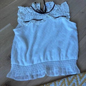 Vit blus med spets och volang - Supersöt vit blus med spets upptill och volangkant nertill. Blusen har ärmlös design, dekorativ knytning i halsen och små diskreta prickar i tyget. Perfekt för dig som gillar romantisk och feminin stil.