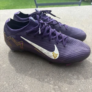 Nike Mercurial Mbappé Elite fotbollsskor lila - Snygga Nike Mercurial fotbollsskor i samarbete med Mbappé. Skorna är lila med guld- och vita detaljer, har snörning och en lätt, syntetisk ovandel. Sulan har rosa dobbar och coola detaljer. Perfekta för dig som vill sticka ut på planen! Ordinarie pris 2999 sek. Inköpta i maj. Tyvärr för små för mig så bara använt ett par gånger. 