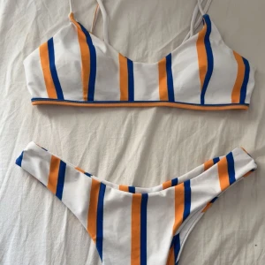 Randig bikini i vitt, orange och blått - Snygg bikini med sportig vibe i vitt med vertikala orangea och blå ränder. Toppen har smala axelband och byxan är högt skuren. Perfekt för stranden eller poolen, och ger en fräsch och färgglad look. Materialet är mjukt och stretchigt syntet. Den har blivit lite missfärgad som syns på bild2, men inget som syns jättemycket 
