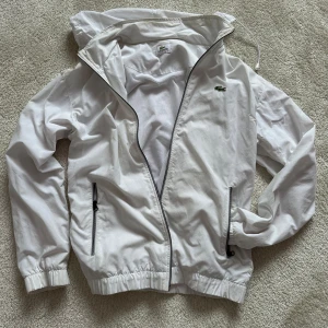Vit vindjacka från Lacoste med huva - Snygg vit vindjacka från Lacoste med dragkedja, huva och två fickor med svarta detaljer. Jackan har elastiska muddar och klassisk krokodil-logga på bröstet. Perfekt för blåsiga dagar och enkel att matcha med allt.