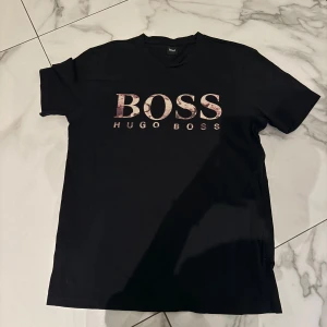 Svart Hugo Boss t-shirt med logga - Svart t-shirt från Hugo Boss med stor BOSS-logga i metallic-rosa och guldiga toner på bröstet. Klassisk passform med rund hals och korta ärmar. Perfekt för dig som gillar stilrena och trendiga plagg med tydlig branding.