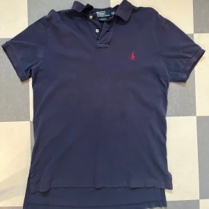 Marinblå pikétröja från Ralph Lauren - Snygg marinblå pikétröja från Ralph Lauren med klassisk krage och två knappar. Tröjan har det ikoniska röda broderade polospelaren på bröstet och är i bomull. Custom fit-modell som sitter riktigt bra och passar perfekt till sommaren. Personen på bilden är 180 cm lång och väger 70 kilo.