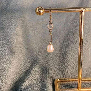 Guldörhänge med pärla och kristall - Snyggt hängande örhänge i guldton med en rund klar kristall och en droppformad vit pärla. Kedjelänk mellan kristallen och pärlan ger en modern touch. Perfekt för dig som gillar stilrena smycken med lite extra detalj.