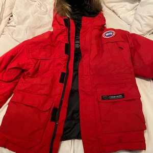 Röd dunjacka från Canada Goose - Säljer en röd Canada Goose dunjacka med huva och pälskant. Jackan har flera stora fickor, klassisk patch på bröstet och dragkedja med kardborre. Perfekt för kalla vinterdagar och riktigt snygg streetstil.