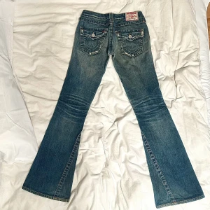 True Religion bootcut jeans blå, stl 24 - Säljer ett par ikoniska True Religion jeans i klassisk blå tvätt med bootcut passform. Snygga kontrastsömmar, broderade bakfickor och slitna detaljer framtill. Jeansen har låg midja och är tillverkade i mjukt denim med cool vintagekänsla. Midjemått: 35.5 cm