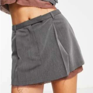 Grå kortkjol med veck - Snygg grå kortkjol med trendiga veck och bälteshällor i midjan. Kjolen har en stilren look och passar perfekt till en cool vardagsoutfit. Materialet känns som polyester och ger en dressad känsla.