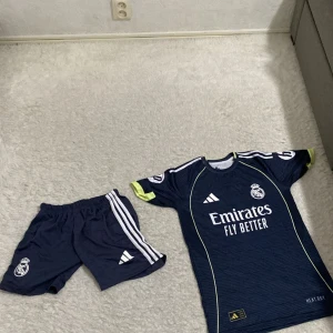 Real Madrid Bellingham matchställ Adidas - Säljer ett Real Madrid matchställ med Bellingham 5 på ryggen. Tröjan är marinblå med tunna ränder, vita och neongula detaljer samt klubbmärke och sponsortryck. Tillverkad i lätt funktionsmaterial (AEROREADY). Korta ärmar och klassisk passform. Shorts i samma stil ingår.