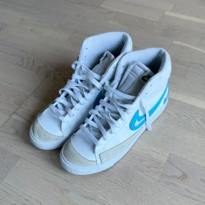 Nike Blazer Mid vit/blå sneakers - Nike Blazer Mid sneakers i vitt skinn med ljusblå swoosh och detaljer. Högt skaft, klassisk snörning och beige mocka vid tån. Snygg retrostil med tydlig Nike-logga på hälen. Perfekta för dig som gillar streetwear och vill sticka ut med färg.