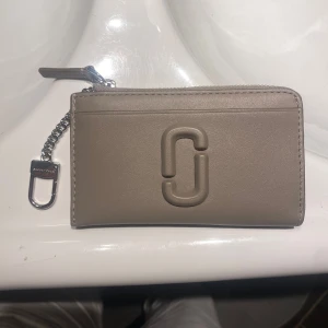 Beige plånbok från Marc Jacobs - Snygg beige plånbok från Marc Jacobs i skinn med silverfärgad kedja och dragkedja. Plånboken har det ikoniska Marc Jacobs-logot på framsidan och ett smidigt fack för kort och mynt. Perfekt att haka på väskan eller använda separat.