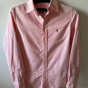 Rosa skjorta från Ralph Lauren  - Snygg rosa skjorta från Ralph Lauren i custom fit. Klassisk design med knappar hela vägen, lång ärm och ikonisk broderad logga på bröstet. Skjortan är gjord i mjuk bomull och passar perfekt till jeans eller chinos.