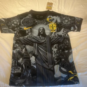 Nike Brasil svart grafisk fotbollströja - Unik svart Brasil-fotbollströja från Nike med grymt detaljerad grafisk print av Kristusstatyn och brasilianska motiv. Gula Nike- och Brasil-loggor på bröstet. Tillverkad i lätt och ventilerande material, perfekt för dig som vill sticka ut på planen.