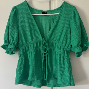 Grön blus med knyt från Gina Tricot - Supersöt grön blus från Gina Tricot med v-ringning och knyt framtill. Blusen har korta puffärmar med volangkant. Tillverkad i ett lätt material som känns luftigt och fräscht. Pris går att diskutera 