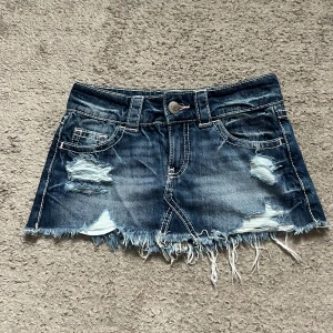 Ripped jeanskjol med fransig kant - Säljer en kort jeanskjol i mörkblå denim med slitna detaljer och fransig nederkant. Kjolen har klassiska fickor fram och bak, bälteshällor och coola slitningar för en edgy vibe. Perfekt för dig som gillar streetstyle och vill sticka ut.