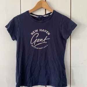Mörkblå Gant t-shirt med tryck - Snygg mörkblå t-shirt från Gant med vit text 'New Haven Gant' på bröstet. Klassisk rund halsringning och korta ärmar. Skön och mjuk bomullskvalitet, perfekt till jeans eller shorts. Enkel och stilren design med sportig vibe.