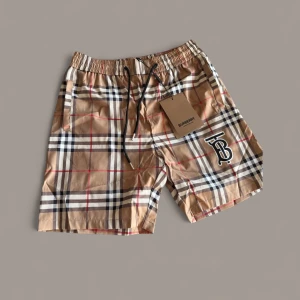 Burberry shorts - Snygga shorts från Burberry i klassiskt beige rutigt mönster med svarta, vita och röda detaljer. Elastisk midja med svart snörning och Burberrys TB-logga framtill. Perfekta för sommaren och ger en lyxig vibe till din outfit.