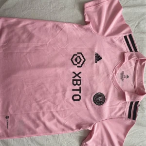 Inter Miami Messi fotbollströja rosa - Rosa Inter Miami CF fotbollströja från Adidas med Messi och nummer 10 på ryggen. Tröjan har svarta detaljer, klubbmärke på bröstet och tre svarta ränder på axeln. Tillverkad i lätt och ventilerande Aeroready-material, perfekt för fotbollsplanen.