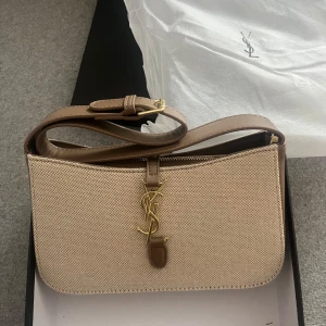 Beige axelväska från Saint Laurent - Snygg beige axelväska från Saint Laurent med klassisk YSL-logga i guld framtill. Väskan är i canvas med detaljer och rem i ljusbrunt skinn. Den har dragkedja upptill och justerbar axelrem. Perfekt storlek för det viktigaste och en riktigt stilren look. Aldrig använd. Har inget äktehetsbevis, därav priset❤️