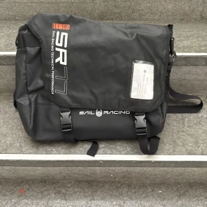 Svart datorväska Sail Racing SR77 - Svart datorväska från Sail Racing SR77 med justerbar axelrem, två spännen framtill och praktiskt fack för namnlapp. Väskan har en sportig look och är tillverkad i slitstarkt syntetmaterial. Perfekt för dig som vill ha en stilren och funktionell väska till plugget eller jobbet.