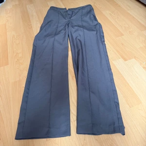 Grå wide kostymbyxor från Madlady - Snygga gr/lila hlgmidjade kostymbyxor från Madlady i modell med vida ben och pressveck. Byxorna har hällor för bälte och stängs med dragkedja och knapp. Perfekta för en clean och stilren look.