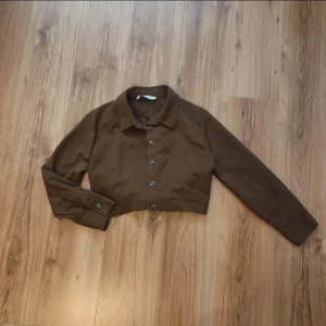Brun croppad overshirt från Zara - Snygg croppad brun overshirt från Zara med klassisk krage och knäppning framtill. Jackan har långa ärmar med knappar vid ärmslut och är tillverkad i ett mjukt bomullstyg. Perfekt för dig som gillar en clean och stilren look. Står att det är storlek L men skulle säga att det är en M