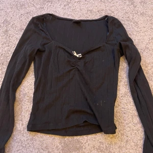 Svart ribbad långärmad topp med rosett - Svart långärmad topp med ribbat tyg och en liten vit rosett framtill vid den v-ringade halsen. Snygg passform som sitter tajt mot kroppen och ger en clean look. Perfekt att styla med jeans eller kjol för en trendig outfit.