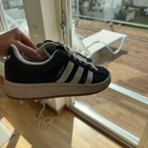 Adidas Campus sneakers - Svarta Adidas Campus sneakers med klassiska vita ränder och logga på hälen. Skorna har vit snörning, rund tå och platt sula. Ovandelen är i mocka och insidan är fodrad i vitt tyg. Perfekt för dig som gillar retrostil och streetwear. Något slitna och smutsiga som säkert går att tvätta bort.