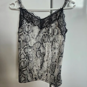 Linnetopp med ormmönster och spets - Snygg linnetopp med svartvitt ormmönster och tunna axelband. Toppen har en cool spetsdetalj längs urringningen som ger extra edge. Perfekt att styla med jeans eller kjol för en trendig look.