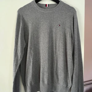 Grå stickad tröja Tommy Hilfiger - Klassisk grå stickad tröja från Tommy Hilfiger med rund halsringning och diskret logga broderad på bröstet. Tröjan har ribbade muddar och är långärmad, perfekt för lager-på-lager. Enkel och stilren design som funkar till allt.