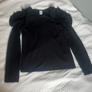 Svart långärmad topp med volang Lindex - Svart långärmad topp från Lindex med coola volangdetaljer på axlarna. Toppens material är mjukt och stretchigt, perfekt för dig som vill sticka ut lite extra. Enkel rund halsringning och stilren look som passar till många outfits.