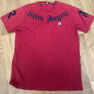 Röd Palm Angels x Moncler t-shirt - Röd t-shirt från Palm Angels och Moncler med svart tryck fram och bak. Stort 'Palm Angels' tryck över bröstet och Moncler-logga broderad på vänster sida. Klassisk rund hals och korta ärmar. Snygg streetwear-vibe och skön bomullskvalitet.