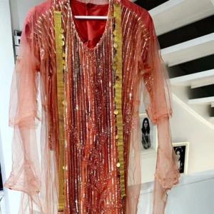 Glittrig rosa kurdisk klänning med guldiga detaljer - Säljer en unik rosa kurdisk klänning med pärlor och glittriga paljetter och guldiga band. Klänningen har långa ärmar, v-ringning och dekorativa detaljer framtill. Perfekt för dig som vill sticka ut med en färgstark och glamorös look. Säljer även guldiga detaljerna vid sidan i första bilden. Storleken Xs och S✨