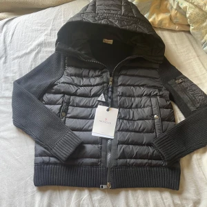 Svart Moncler cardigan - Svart cardigan från Moncler med stickade ärmar och ribbade muddar. Jackan har huva, dragkedja framtill och två fickor med tryckknappar. Materialmix av polyester och stickat ger en snygg kontrast. Perfekt för kyliga dagar och streetstyle. Använd 1 gång eftersom den inte passade mig alla scans funkar på den och inga defekter 