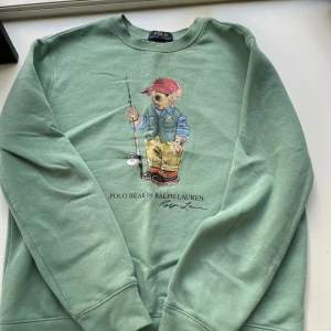 Grön Polo Bear tröja från Ralph Lauren - Snygg mintgrön sweatshirt från Polo Ralph Lauren med ikoniskt Polo Bear-tryck framtill. Tröjan har rund hals, långa ärmar och är tillverkad i mjuk bomulls- och polyestermix. Perfekt för dig som gillar streetwear med en preppy twist.