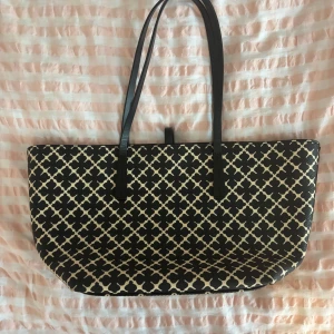 Svartvit shoppingväska från By Malene Birger - Stilren shoppingväska från By Malene Birger i svart och vitt med klassiskt grafiskt mönster. Väskan har långa svarta handtag, rymligt beige innerfoder och stängs med ett smidigt spänne. Perfekt för dig som vill ha plats för allt du behöver under dagen. 