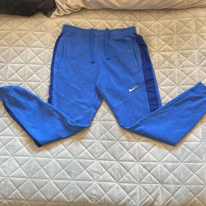 Blå mjukisbyxor från Nike - Snygga blå mjukisbyxor från Nike med mörkblå sidopaneler och klassisk logga på benet. Byxorna har dragsko i midjan, ribbade muddar och är gjorda i mjukt material som känns skönt mot huden. Perfekta för chill eller träning.
