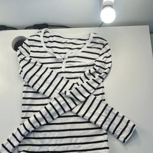 Randig långärmad tröja från H&M - Säljer en vit och svart långärmad tröja från H&M med klassiskt randigt mönster. Tröjan har rund halsringning och knappar framtill. Tillverkad i mjuk bomull, perfekt för en chill och stilren look.