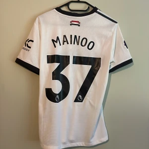 Manchester United Mainoo #37 Adidas tröja - Säljer en Manchester United Mainoo #37 fotbollströja från Adidas. Tröjan är vit med svarta och röda detaljer, svarta ränder på axlarna och tryckta loggor från Premier League och Snapdragon. Tillverkad i ventilerande meshmaterial, kortärmad och med klassisk rund hals.