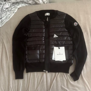 Moncler cardigan  - Snygg svart cardigan från Moncler med quiltad framsida och två fickor med knapp. Cardigan har dragkedja framtill och Moncler-logga på ärmen. Materialmix med dun och stickade partier ger en modern look. Perfekt för lager-på-lager-stil.