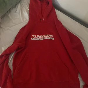 Röd JLindeberg hoodie Black Piste Tour - Röd hoodie från JLindeberg med tryck 'Black Piste Tour 2018-19' på bröstet och blå siluetter på ryggen. Klassisk känguruficka och huva med snörhål. Perfekt statement-plagg för dig som gillar streetwear och sportig stil.