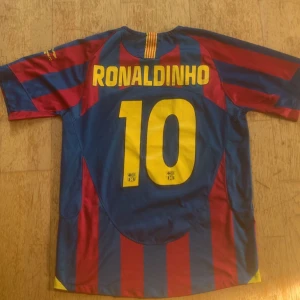 FC Barcelona Ronaldinho 10 tröja Nike - Säljer en klassisk FC Barcelona fotbollströja med Ronaldinho och nummer 10 på ryggen. Tröjan är randig i blått och rött med gula detaljer, klubbmärke på bröstet och Nike-logga. Tillverkad i lätt och ventilerande material, perfekt för dig som älskar fotboll och Barca.