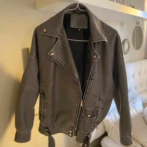 Svart bikerjacka med bälte, stl S - Säljer en svart bikerjacka i skinnimitation med coola detaljer som sned dragkedja, silvriga knappar och bälte i midjan. Jackan har lång ärm och markerad krage, perfekt för en edgy streetstyle-look. Perfekt nu inför hösten 🍂😍🤍