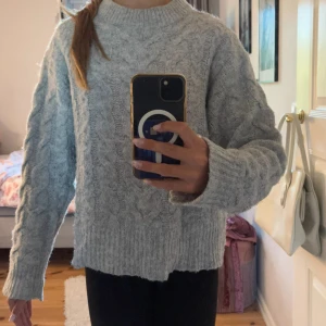 Grå stickad tröja från Gina Tricot - Mysig grå stickad tröja från Gina Tricot med kabelstickat mönster. Tröjan har en hög, ribbad halsringning och långa ärmar med breda muddar. Perfekt för lager-på-lager och chill dagar. Snygg loose fit som ger en avslappnad vibe.