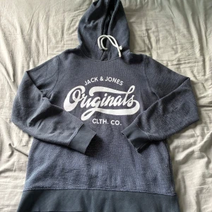 Mörkgrå hoodie Jack & Jones Originals storlek M - Snygg mörkgrå hoodie från Jack & Jones Originals med vit logga framtill. Tröjan har huva med vit snörning och breda muddar vid ärmslut och nederkant. Perfekt för en chill och avslappnad stil.