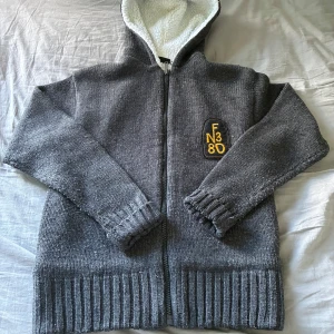 Grå stickad jacka med huva storlek M - Mysig grå stickad teddyjacka med huva och hel dragkedja. Insidan är fodrad med vit teddyfleece för extra värme. Jackan har ribbade muddar och nederkant samt coola gula patchar på bröstet och ena ärmen. Perfekt för kyliga dagar.