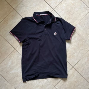 Svart Moncler pikétröja med röd/vit kant - Svart pikétröja från Moncler med klassisk krage och korta ärmar. Tröjan har röda och vita detaljer på krage och ärmslut samt Moncler-logga på bröstet. Tillverkad i bomull för en skön och stilren look. Storlek medium. Pris kan diskuteras vid snabbaffär. 