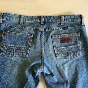 Wrangler blå jeans bootcut - Klassiska blå jeans från Wrangler med bootcut passform, modellen heter Megan. Snygga kontrastsömmar och ikoniska W-detaljer på bakfickorna. Jeansen har låg midja, fem fickor och är tillverkade i slitstarkt bomullsjeans. Perfekta för dig som gillar en tidlös denimlook. Tyvärr är de slitna längst ner vid hälen, samt lite slitna i sömmarna som visas på bilden. Innerbenslängd ca. 82 cm. Midjemått rakt över ca. 40 cm. Skriv innan köp och för fler bilder!!☺️