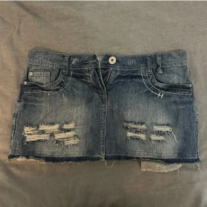 Mörk jeanskjol med slitningar - Snygg mörkblå jeanskjol med slitna detaljer framtill och bakfickor med dekorativa sömmar. Kjolen har rå kant nertill och klassiska jeansfickor. Perfekt för en cool och avslappnad stil.
